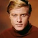 Foto in primo piano di Robert Redford che indossa un maglione marrone a collo alto su sfondo marrone più chiaro